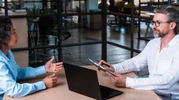 Dos personas conversando en una oficina, una con portátil y otra con tableta.